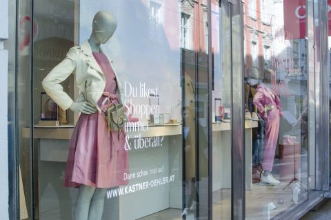 Schaufenster mit einer Schaufensterpuppe in einem rosa Kleid und einer leichten Jacke. Im Glasfenster steht ein deutscher Werbetext für den Einkauf. Eine Straßenspiegelung zeigt verschwommene Passanten und Stadtgebäude.