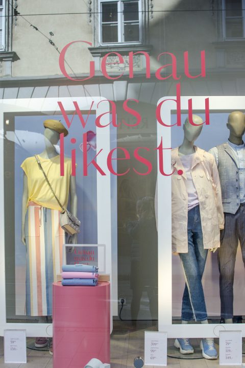 In einem Schaufenster sind Schaufensterpuppen in legerer Kleidung zu sehen, darunter ein gestreifter Rock mit einem gelben Oberteil und eine leichte Jacke mit Jeans. Der Text im Fenster lautet: „Genau was du liebst“, was auf Deutsch „Genau das, was du magst“ bedeutet.