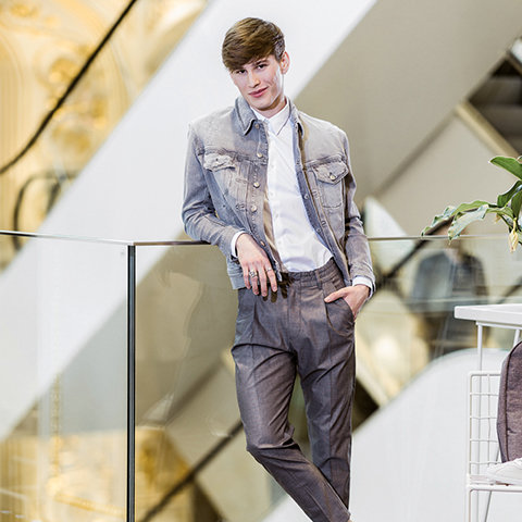 Eine Person in heller Jeansjacke und grauer Hose lehnt lässig an einem Glasgeländer in einem modernen, hell erleuchteten Raum. Die Hände hat sie in den Taschen, die Haare sind kurz und braun.