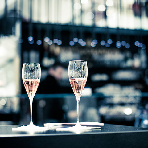 Zwei Gläser Rosé-Champagner auf einem dunklen Tisch. Der Hintergrund ist leicht verschwommen und zeigt eine schwach beleuchtete Bar mit dezentem Bokeh-Licht. Die Szene vermittelt ein Gefühl von Eleganz und Feierlichkeit.