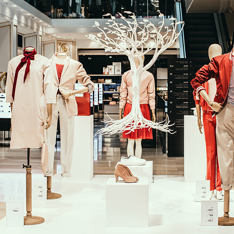 Eine Verkaufsausstellung mit Schaufensterpuppen in modischer Kleidung, darunter rot-weiße Outfits. Zwischen ihnen steht eine dekorative weiße Baumskulptur, auf weißen Podesten sind verschiedene Schuhe ausgestellt. Im Hintergrund ist eine Rolltreppe zu sehen.