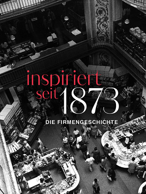 Schwarzweißfoto eines belebten Kaufhauses mit geschäftigen Menschenmengen. Der rote Text lautet „inspiriert seit 1873“ und der weiße Text „DIE FIRMENGESCHICHTE“, was auf eine Ausstellung zur Firmengeschichte hinweist.