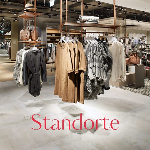Ein schickes Interieur eines Bekleidungsgeschäfts mit hängenden Kleidungsstücken in neutralen Tönen wie Beige und Grau. Das Geschäft bietet eine Vielzahl von Modeartikeln und Accessoires. Das Wort „Standorte“ ist deutlich auf dem Boden zu sehen.