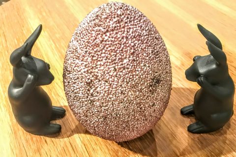 Zwei kleine schwarze Kaninchenskulpturen sitzen einander gegenüber auf einer Holzoberfläche, zwischen ihnen befindet sich ein großes strukturiertes, glitzerndes Ei.