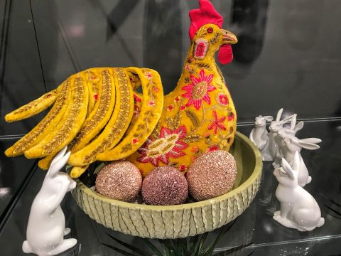 Eine bunt bestickte Henne sitzt in einer dekorativen Schale, umgeben von glitzernden Eiern. Vier weiße Keramikhasen sind um die Schale herum angeordnet und sorgen für eine verspielte Szene.