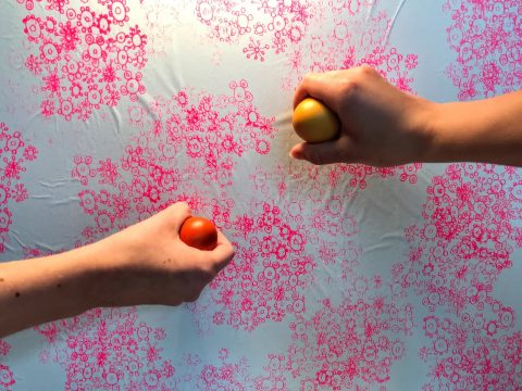 Zwei Hände halten rote und gelbe Eier vor einem weiß-rosa Hintergrund mit Blumenmuster. Die Hände sind so positioniert, als würden sie die Eier aneinanderschlagen.