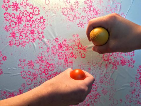 Zwei Hände drücken gelbe und orangefarbene Bälle gegen eine Wand und erzeugen so rosafarbene und orangefarbene Blumenmuster auf einer weißen Oberfläche.