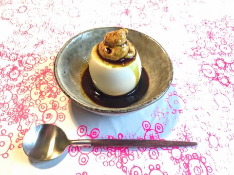 Ein kleines Dessert auf einem grauen Teller besteht aus einem cremigen Pudding mit Soße und einem Klecks Karamell oder Schlagsahne, platziert auf einer Tischdecke mit Blumenmuster. Neben dem Teller liegt ein Edelstahllöffel.