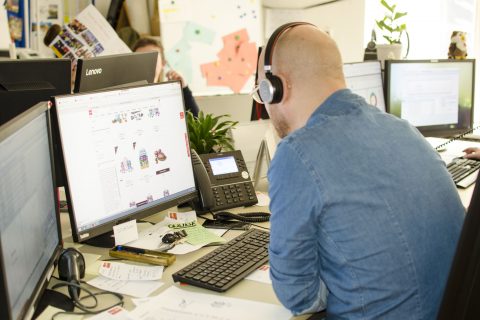 Eine Person mit Kopfhörern sitzt an einem Schreibtisch und arbeitet an einem Computer mit mehreren Monitoren. Auf dem Schreibtisch liegen Papiere, ein Telefon und Büromaterial herum. Im Hintergrund ist eine Pflanze zu sehen und an der Wand hängt eine bunte Karte.