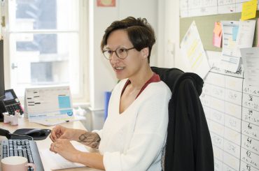 Eine Person mit kurzen Haaren und Brille sitzt lächelnd an einem Schreibtisch. Sie trägt einen weißen Pullover und über dem Stuhl hängt eine schwarze Jacke. Hinter ihr hängen ein schwarzes Brett und ein Kalender an der Wand. Auf dem Schreibtisch liegen Papiere und ein Telefon.