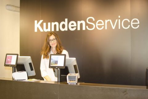 Eine Frau mit Brille steht an einem Kundenservice-Schalter mit der Aufschrift "KundenService". Sie sitzt hinter zwei Computermonitoren und lächelt in die Kamera. Das Ambiente scheint ein moderner, einladender Servicebereich zu sein.