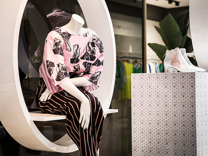Schaufensterpuppe in runder Vitrine, trägt ein rosa-schwarz gemustertes Oberteil und gestreifte Hose. Daneben steht ein Podest mit weißen Stöckelschuhen. Modernes, stilvolles Interieur mit Pflanzen im Hintergrund.