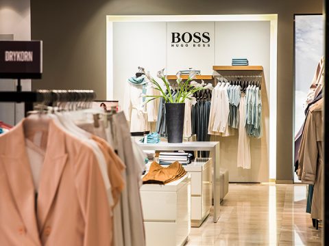 Inneneinrichtung eines Bekleidungsgeschäfts mit Abteilungen von Hugo Boss und Drykorn. Die Auslage umfasst ordentlich angeordnete Hemden, Jacken und eine große schwarze Vase mit weißen Blumen. Neutrale und Pastellfarben dominieren die Bekleidungsauswahl.