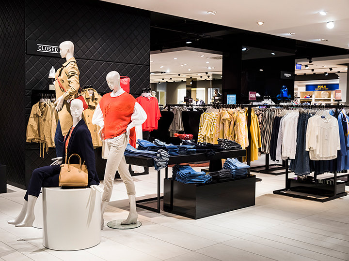 Innenansicht eines Bekleidungsgeschäfts mit Schaufensterpuppen in modischen Outfits. Kleiderständer mit verschiedenen Kleidungsstücken, darunter Jacken, Hemden und Hosen, sind ordentlich angeordnet. Gefaltete Jeans werden auf einem Tisch in der Nähe unter heller Ladenbeleuchtung ausgestellt.