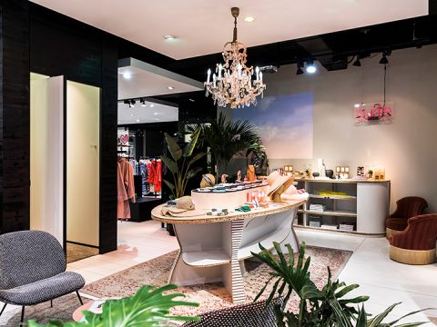 Ein stilvolles Boutique-Interieur mit einem zentralen Ausstellungstisch für Accessoires. Darüber hängt ein Kronleuchter, und Kleiderständer säumen die Wände. Man sieht einen gemütlichen Sitzbereich mit modernen Stühlen, und Grünpflanzen sorgen für eine frische Note.