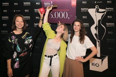 Drei Frauen stehen vor der Kulisse eines Modepreises. Die Frau in der Mitte hält voller Begeisterung einen Preisscheck über 5.000 € hoch. Sie alle lächeln und tragen modische Kleidung und feiern ihren Erfolg.