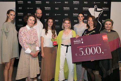Eine Gruppe von acht Personen steht lächelnd zusammen, zwei Frauen halten einen großen Scheck über 5.000 Euro in den Händen. Sie posieren vor einem Hintergrund mit der Silhouette einer Schaufensterpuppe und Logos von Modepreisen.