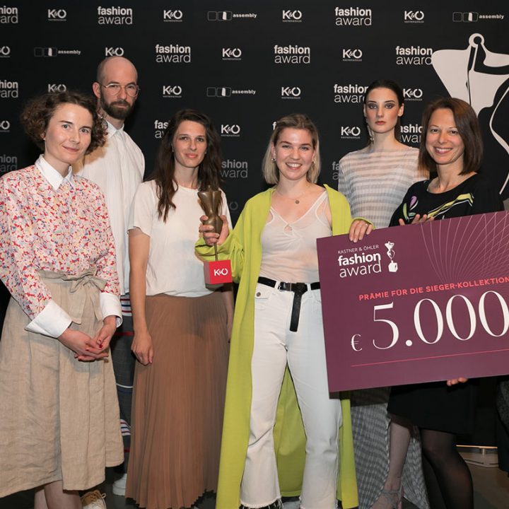 Eine Gruppe von acht Personen posiert bei einer Modeveranstaltung. Einer hält einen großen Preisscheck über 5.000 Euro in der Hand. Sie stehen vor einem schwarzen Hintergrund mit Logos von Modeveranstaltungen. Einige tragen stilvolle, moderne Outfits.