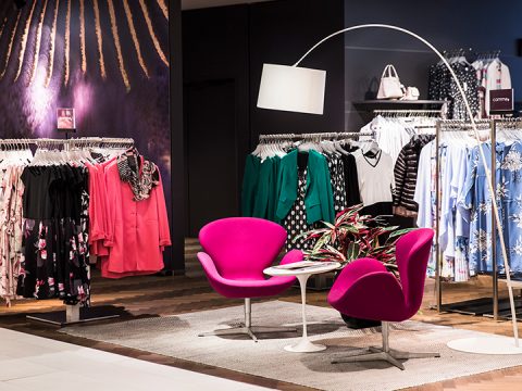 Ein Bekleidungsgeschäft mit Regalen voller bunter Kleidungsstücke. Zwei leuchtend rosa Stühle stehen einander um einen kleinen weißen Tisch herum gegenüber, unter einer modernen gewölbten Stehlampe. Verschiedene Outfits, darunter Kleider und Blusen, sind ordentlich angeordnet.