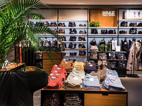 Eine Bekleidungsgeschäft-Auslage mit ordentlich gefalteten Hosen in verschiedenen Farben auf einem zentralen Tisch. Regale im Hintergrund sind mit weiteren Kleidungsstücken gefüllt. Im Vordergrund ist eine große Pflanze teilweise sichtbar. Das Schild „BRAX Feel Good“ befindet sich an der Rückwand.