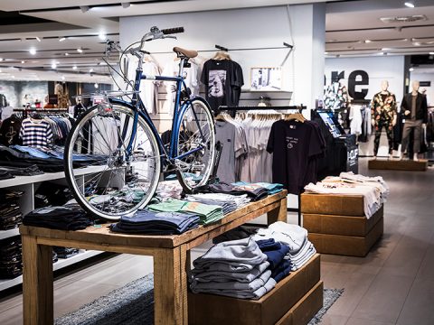 Innenansicht eines Einzelhandelsgeschäfts mit einem Holztisch, auf dem gefaltete Hemden ausgestellt sind. Auf dem Tisch steht ein blaues Fahrrad. Im Laden sind Kleidungsstücke und Schaufensterpuppen in modischen Outfits angeordnet, was eine trendige Atmosphäre schafft.