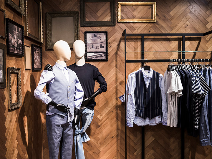 Ein Schaufenster in einem Boutique-Kleidergeschäft mit zwei Schaufensterpuppen. Eine trägt eine hellblaue Weste und karierte Hosen, die andere einen schwarzen Pullover und blaue Hosen. In der Nähe steht ein Ständer mit Hemden und Westen. Verschiedene Bilderrahmen schmücken die hölzerne Wand im Fischgrätenmuster.