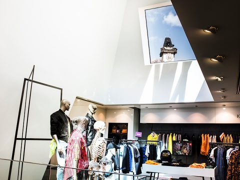 Ein hell erleuchtetes Bekleidungsgeschäft mit mehreren Schaufensterpuppen in farbenfrohen Outfits. An der Decke befindet sich ein großes Oberlicht, durch das man vor einem blauen Himmel ein Stück eines Uhrturms sehen kann. Im Hintergrund sind reihenweise Kleidungsstücke und Accessoires ordentlich ausgestellt.