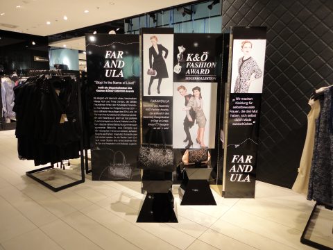 In einem Modegeschäft sind Schaufensterpuppen in eleganten Outfits und Accessoires zu sehen. Die Einrichtung umfasst Tafeln mit Texten zu Modepreisen und Slogans, die vor einem modernen Ladeninterieur mit Kleiderständern und stilvollem Dekor stehen.
