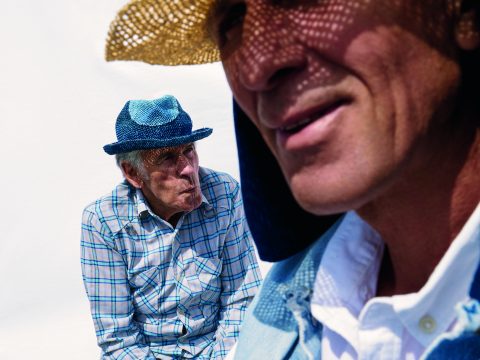 Zwei Männer mit Hüten. Der Mann im Vordergrund trägt einen geflochtenen Strohhut und ein gemustertes Hemd und lächelt. Der Mann im Hintergrund sitzt in einem karierten Hemd und schaut weg. Er trägt einen blauen Hut. Der Hintergrund ist schlicht weiß.