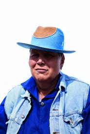 Eine Person mit einem blau-braunen Hut, einer Jeansweste und einem blauen Hemd blickt vor einem schlichten Hintergrund in die Kamera.