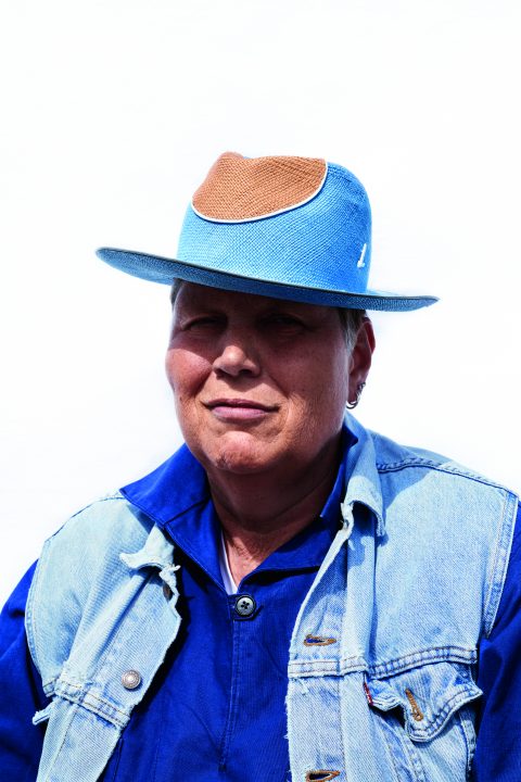 Eine Person mit einem blau-braunen Hut, einer Jeansweste und einem blauen Hemd blickt vor einem schlichten Hintergrund in die Kamera.