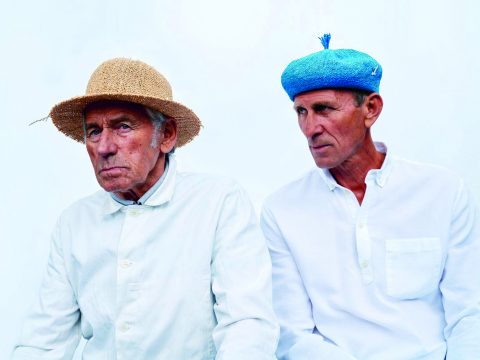 Vor einem schlichten Hintergrund sitzen zwei ältere Männer in weißen Hemden und Hüten. Einer trägt einen Strohhut, der andere ein blaues Barett mit blauer Quaste.