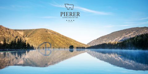 In einem ruhigen Außenpool spiegelt sich der klare blaue Himmel und die umliegenden bewaldeten Berge. Leichter Nebel steigt aus dem Wasser auf, unter einem Logo mit der Aufschrift „Pierer Alpinwellness seit 1893“. Die Szene strahlt Ruhe und natürliche Schönheit aus.