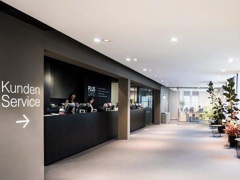 Ein moderner Kundenservicebereich mit einem schwarzen Tresen mit der Aufschrift „Kundenservice“. Hinter dem Tresen steht ein Mitarbeiter mit Computern. Der Raum ist gut beleuchtet und mit Pflanzen dekoriert.