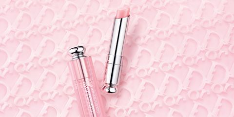 Ein rosa Lippenbalsam auf einem rosa Hintergrund mit geprägten Buchstaben. Der Lippenbalsam hat eine transparente rosa Kappe und eine silberne Tube. Die Buchstaben bilden ein sich wiederholendes Muster und verleihen dem Hintergrund Struktur.