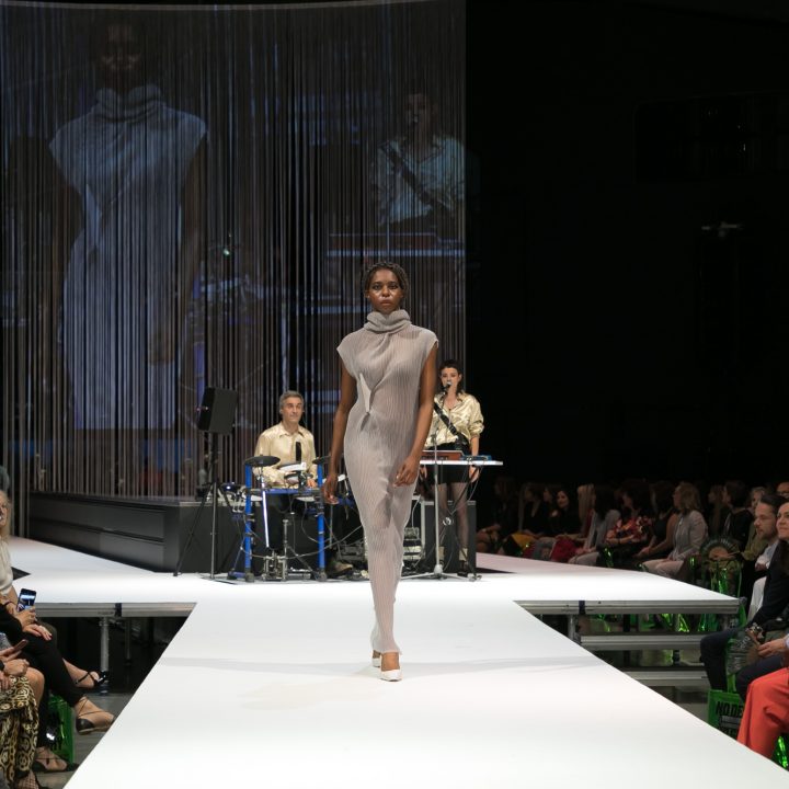 Ein Model läuft in einem grauen Outfit mit hohem Kragen über einen hell erleuchteten Laufsteg. Im Hintergrund tritt eine Band mit einem Sänger und einem Keyboarder auf. Das Publikum sitzt auf beiden Seiten der Bühne und schaut sich die Show an.