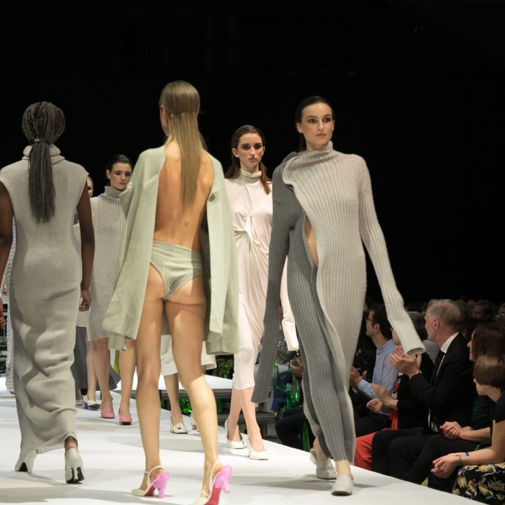 Models laufen in verschiedenen hellen, avantgardistischen Outfits, darunter lange Kleider und Strickwaren, über einen Laufsteg, während auf beiden Seiten des Laufstegs das Publikum sitzt.