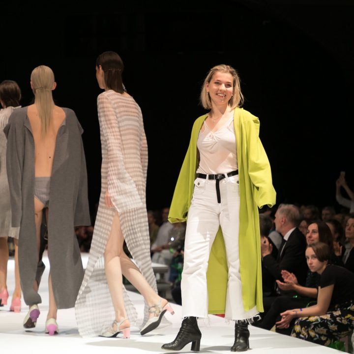 Eine Gruppe von Models läuft in hellen, neutralen Outfits über den Laufsteg. Eine Frau in einem hellgrünen Mantel und weißen Hosen folgt ihnen und lächelt dem applaudierenden Publikum zu. Unter der Bühne schauen die Leute aufmerksam zu.