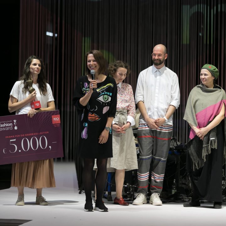 Eine Gruppe von sechs Personen steht auf einer Bühne. Eine Frau hält einen großen Scheck über 5.000 Euro in der Hand. Eine andere Frau spricht in ein Mikrofon. Die anderen lächeln. Alle tragen farbenfrohe, legere Kleidung. Zuschauer beobachten das Geschehen von der Seite.
