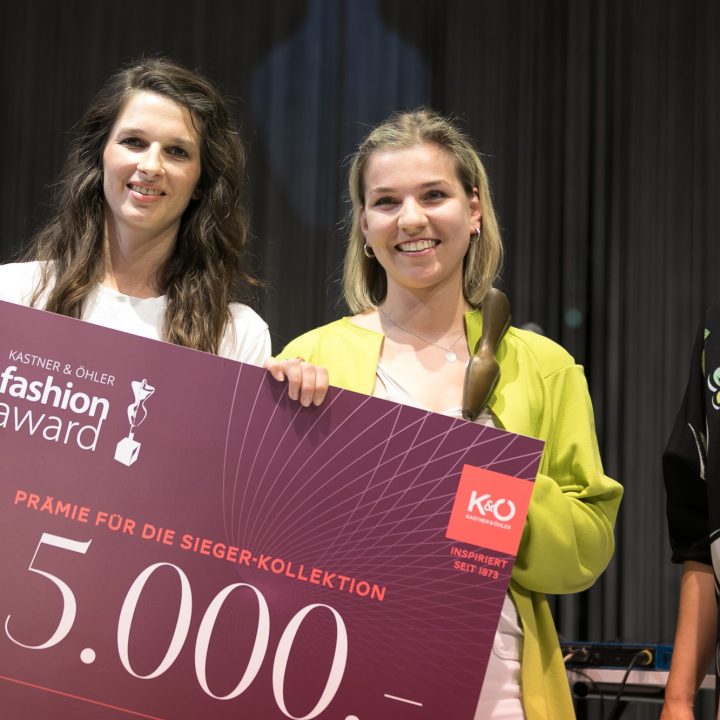 Drei Frauen stehen lächelnd nebeneinander. Die Frau in der Mitte hält einen großen Scheck über 5.000 Euro mit der Aufschrift „Kastner & Öhler Fashion Award“. Sie sind modisch gekleidet und scheinen auf einer Veranstaltung oder Zeremonie zu sein.