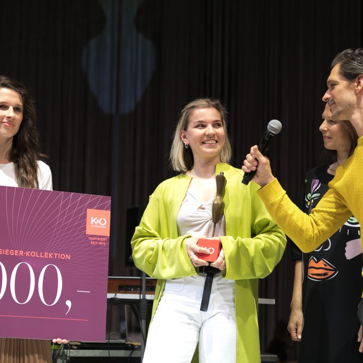 Eine Gruppe von drei Frauen und einem Mann steht auf der Bühne. Eine Frau hält einen großen Scheck über 5.000 Euro und eine Trophäe in der Hand. Der Mann mit dem Mikrofon spricht. Sie scheinen bei einer Preisverleihung zu sein, auf dem Scheck steht „Fashion Award“.