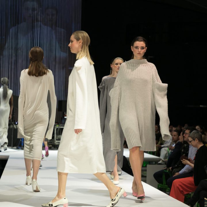 Models laufen in übergroßen, minimalistischen Strickkleidern in neutralen Tönen über den Laufsteg. Das Publikum schaut in einem schwach beleuchteten Saal von beiden Seiten zu, im Hintergrund ist auf einer großen Leinwand der Laufsteg zu sehen.