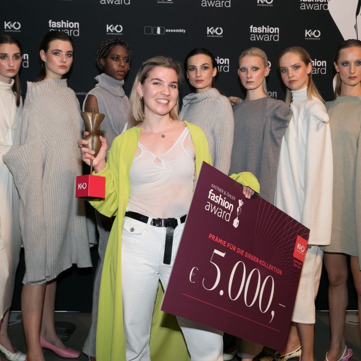 Eine lächelnde Frau hält eine Trophäe und einen großen Scheck über 5.000 Euro in den Händen und steht vor einer Gruppe von acht Models in Kleidern in neutralen Farben. Im Hintergrund sind Logos einer Modepreisverleihung zu sehen.