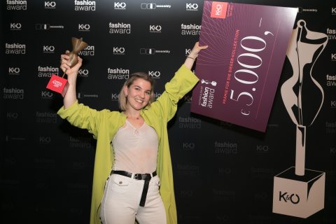 Eine Person in einem hellgrünen Mantel hält bei einer Modepreisverleihung eine Trophäe und einen großen Scheck über 5.000 Euro in den Händen. Im Hintergrund sind die Logos der Veranstaltung und des Sponsors zu sehen. Sie lächeln und wirken aufgeregt.
