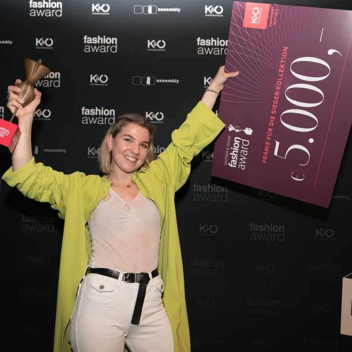 Eine Person in einem hellgrünen Mantel hält bei einer Modepreisverleihung eine Trophäe und einen großen Scheck über 5.000 Euro in den Händen. Im Hintergrund sind die Logos der Veranstaltung und des Sponsors zu sehen. Sie lächeln und wirken aufgeregt.