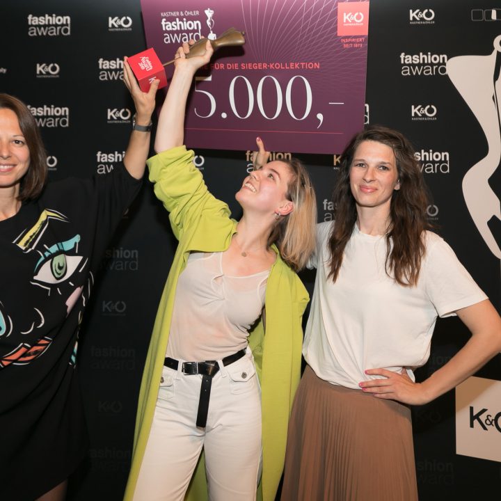 Drei Personen posieren fröhlich vor einem schwarzen Hintergrund mit dem Text „Fashion Award“. Eine Person hält eine rote Award-Karte und eine große Karte mit der Aufschrift „5.000,-“. Sie alle tragen stylische Outfits und strahlen Begeisterung und Feierstimmung aus.