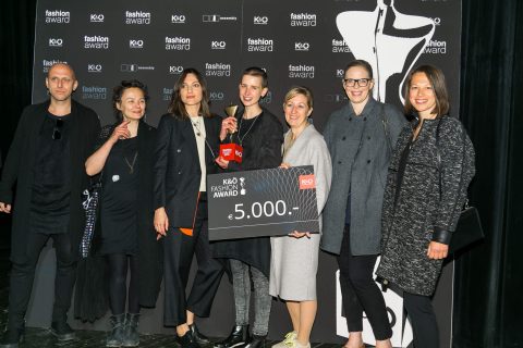 Eine Gruppe von sieben Personen steht lächelnd zusammen. Eine Person hält einen großen Preisscheck über 5.000 Euro in den Händen. Sie stehen auf einer Bühne mit einem schwarzen Hintergrund, auf dem die Logos eines Modepreises zu sehen sind.