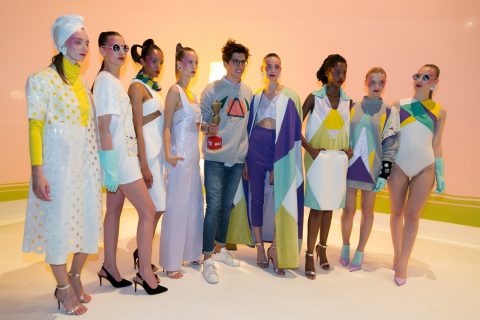 Eine Gruppe von Models in farbenfrohen, geometrisch gemusterten Outfits steht neben einer leger gekleideten Person, die einen roten Becher mit Limonade hält. Die Models tragen verschiedene Kleidungsstile, darunter Kleider und Bodysuits, vor einem pastellfarbenen Hintergrund.