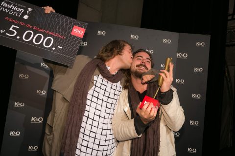 Zwei Menschen feiern bei einer Veranstaltung. Einer hält einen großen Scheck über 5.000 Euro und küsst den anderen auf die Wange, der einen Pokal und eine rote Karte hält. Sie stehen vor einem schwarzen Hintergrund mit einem sich wiederholenden Logomuster.