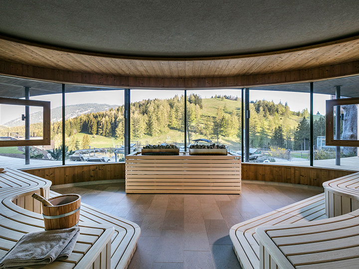 Ein runder Saunaraum mit Holzbänken und großen Panoramafenstern mit Blick auf einen grünen, bewaldeten Hügel. Der Innenraum ist mit natürlichem Licht durchflutet und auf einer der Bänke steht ein Holzeimer.